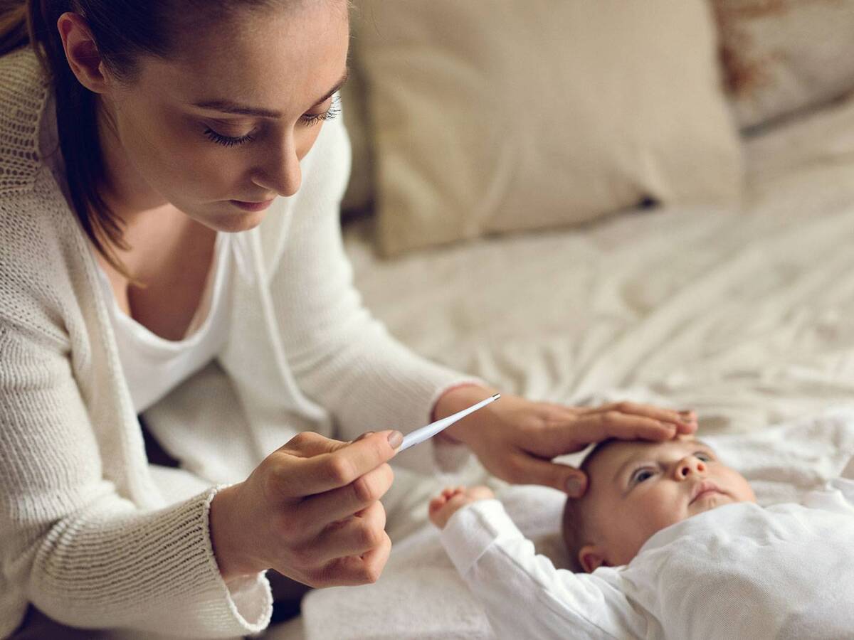 A woman reads a thermometer while touching an infants forehead.