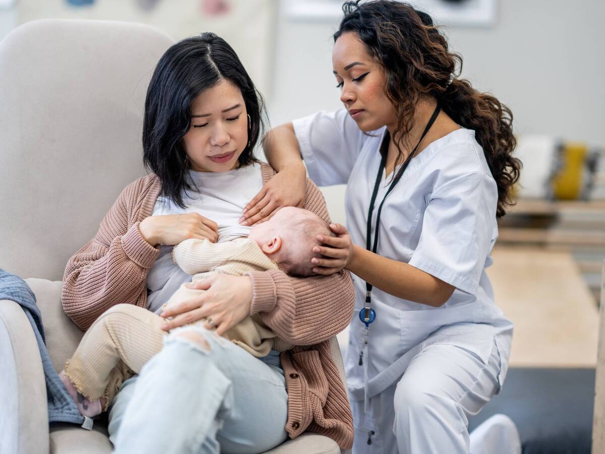 A woman breastfeeding a baby, while being supported and assisted by a woman in medical scrubs.
