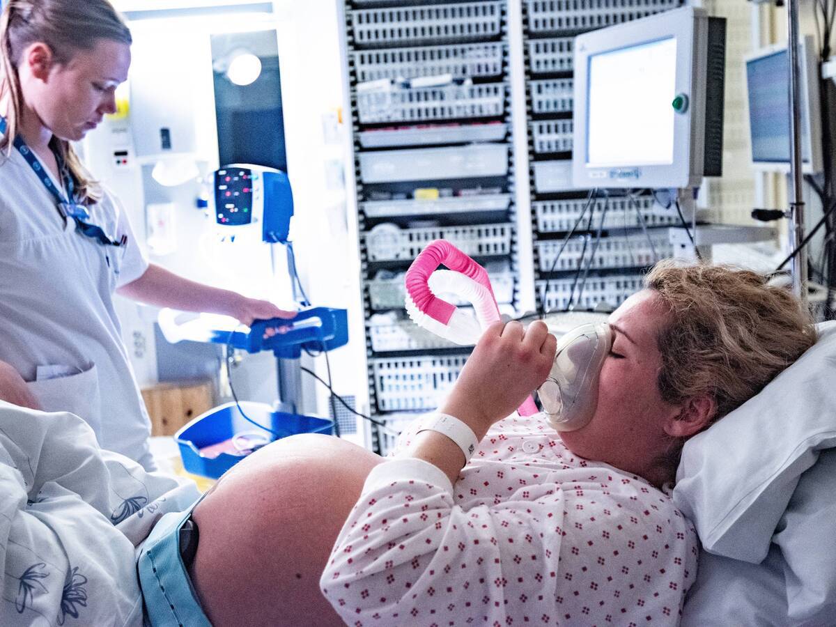 A pregnant woman breathing into an oxygen mask while a nurse arranges medical equipment.