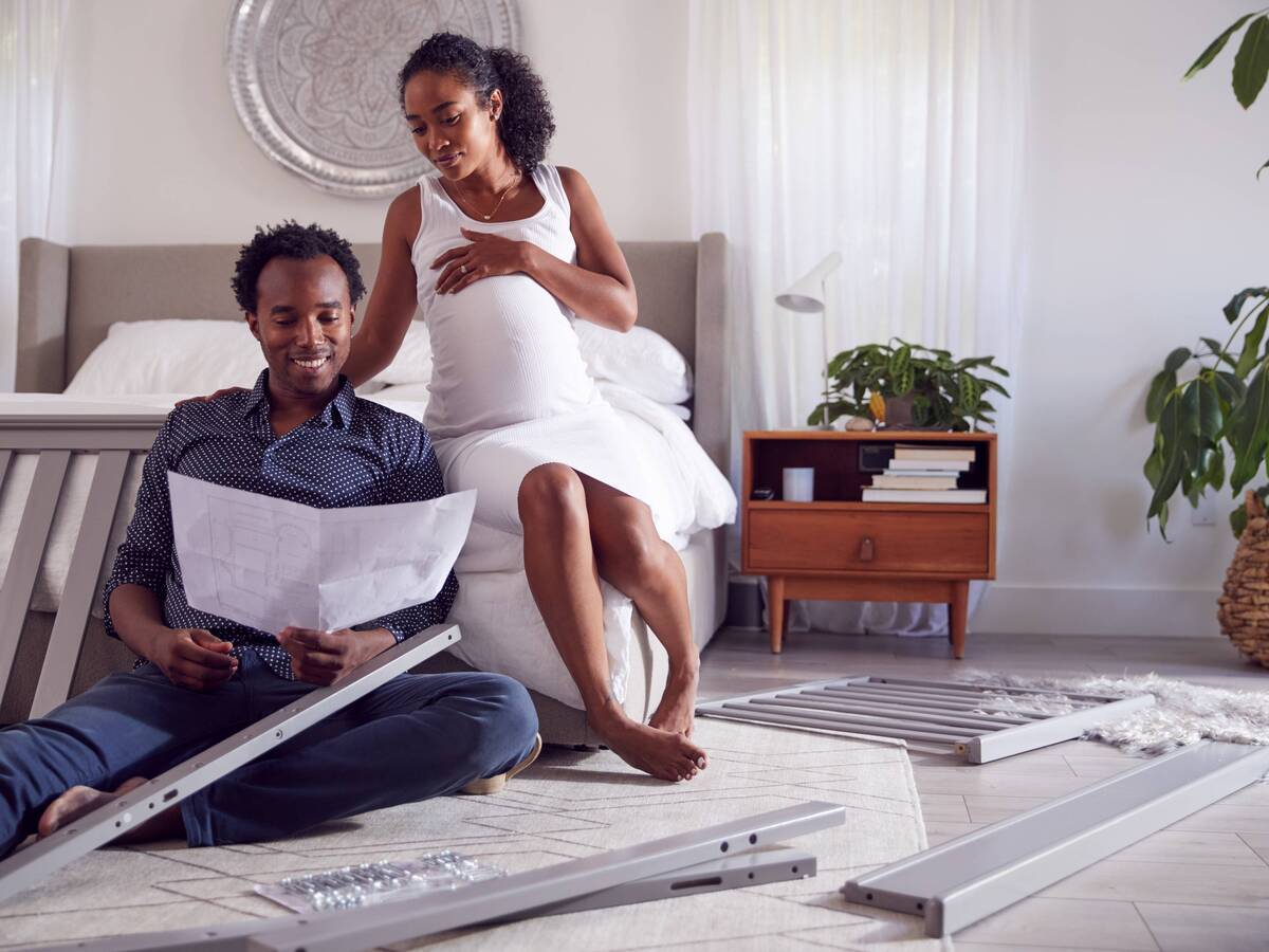 A pregnant woman sitting on the edge of a bed, watching her partner assemble a crib.