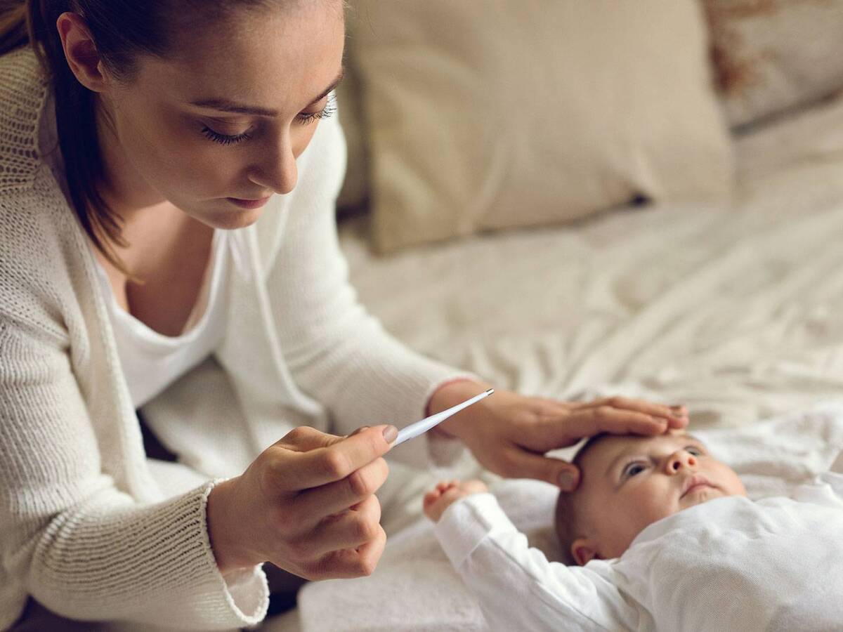 A woman reads a thermometer while touching an infants forehead.
